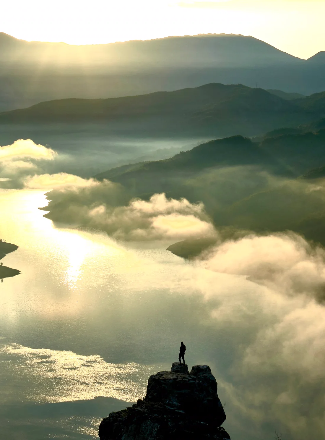 A person standing on a rock above a misty river valley at sunrise