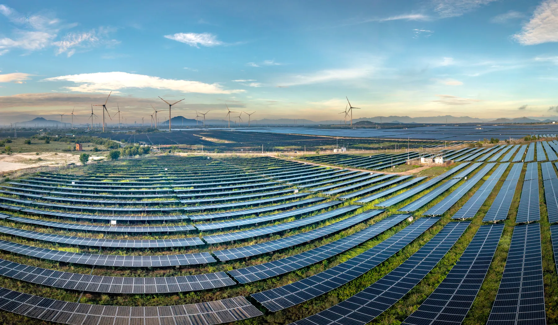 Aerial view of a large solar farm with wind turbines and hills on the horizon